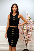 Woman wearing a black outfit with gold buttons standing next to a decorative plant indoors.